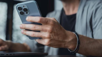 Muž držící iPhone s modrým krytem, sedící u stolu s notebookem v pozadí.