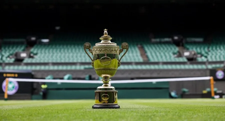Zlatý pohár pro vítěze Wimbledonu stojící na zeleném trávníku hlavního kurtu. Pozadí zobrazuje prázdné tribuny, které podtrhují prestižní atmosféru tenisového turnaje.