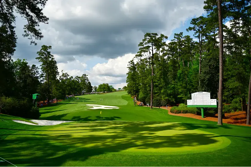 Pohled na ikonické hřiště Augusta National Golf Club během The Masters, lemované stromy a svěže zeleným trávníkem.
