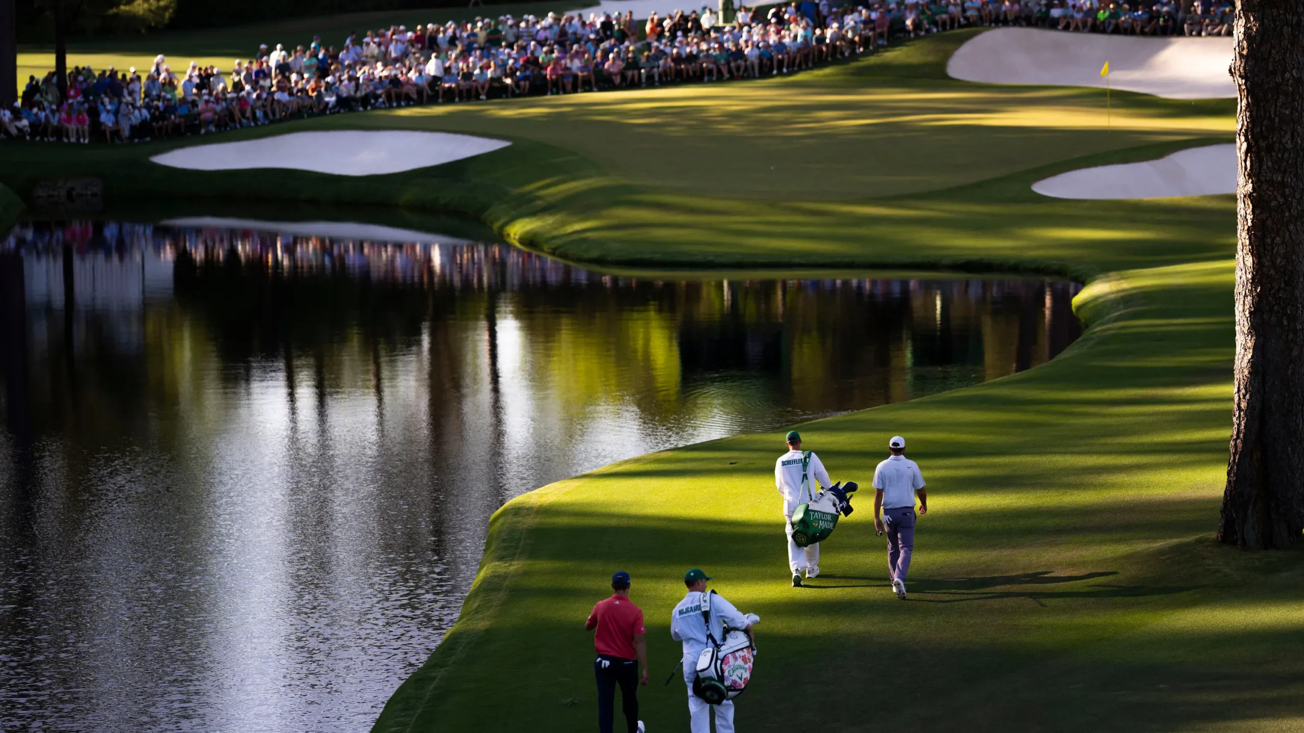 Golfisté a jejich caddies kráčejí podél vodní překážky na jednom z nejslavnějších míst hřiště Augusta během The Masters.