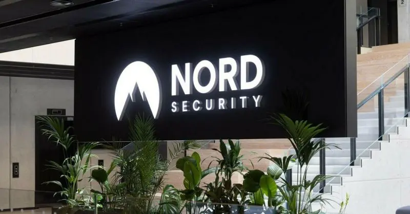 NordSecurity | BestSafeVPN.com Logo společnosti Nord Security na moderní recepci s okolní zelení a schodištěm v pozadí.