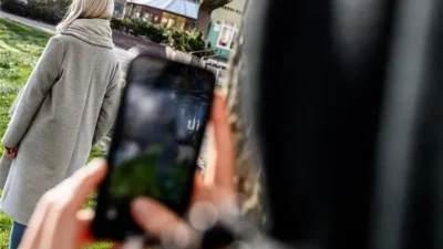 Osoba v šedém kabátu odchází po parku, zatímco ji někdo tajně fotografuje mobilním telefonem ze zálohy. Ilustrace sledování a narušení soukromí.