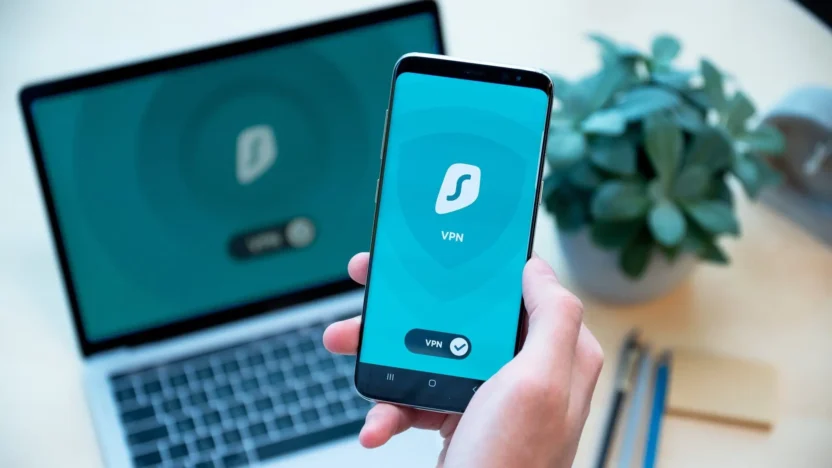 Zapnutá aplikace Surfshark VPN na mobilním telefonu v ruce, v pozadí otevřený notebook s logem Surfshark, pracovní stůl s květinou a tužkami.