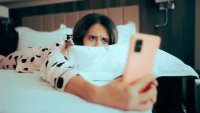 Eine Frau liegt im Bett, schaut besorgt auf ihr Smartphone und wirkt angespannt – Symbolbild für digitale Überwachung, Stalking oder Verletzung der Privatsphäre.